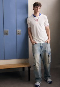 Biała koszulka sportowa z niebieskimi akcentami i logo, w zestawie z jasnoniebieskimi dżinsami cargo z bocznymi kieszeniami oraz białymi trampkami.
