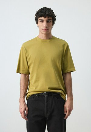 Młody mężczyzna o kręconych ciemnych włosach, ubrany w musztardowozielony T-shirt z krótkim rękawem oraz czarne jeansy z czarnym paskiem, stoi na tle jednolitego białego tła.