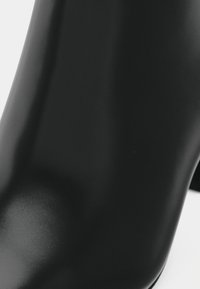 Botim preto até ao tornozelo com um acabamento elegante e brilhante. Apresenta uma forma afinada e um salto alto, destacando a textura suave e detalhes de design mínimos.