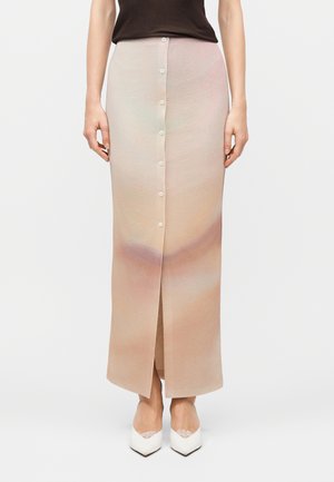 Kobieta ubrana w długą, beżową spódnicę zapinaną na guziki z delikatnym gradientem, zestawioną z ciemną bluzką i białymi butami w szpic z koronkowym wykończeniem.