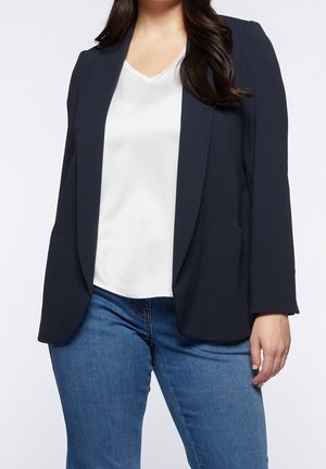 Kobieta ubrana w ciemnogranatową marynarkę na białej bluzce z dekoltem w serek i niebieskie dżinsy, stojąca na jasnym, jednolitym tle.