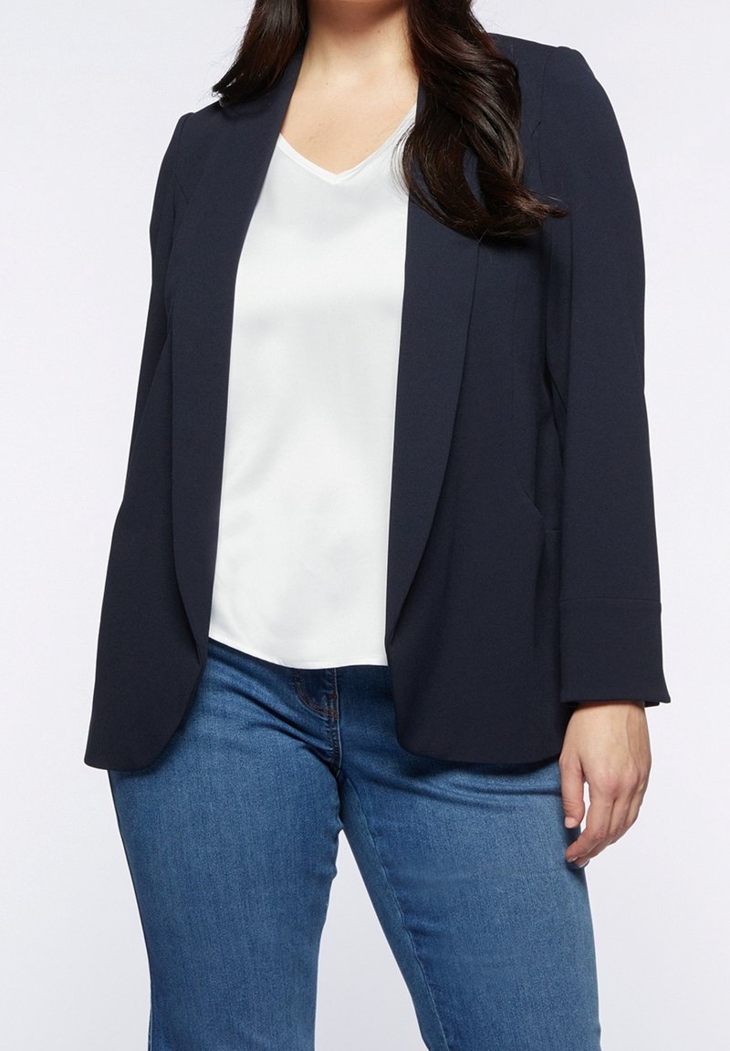 Femme portant un blazer bleu marine foncé sur un chemisier blanc à col en V et un jean denim bleu, debout devant un fond clair uni.