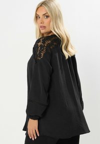 Schwarze Bluse mit Spitzenverzierungen an den Schultern, Dreiviertelärmel mit gerafften Manschetten und strukturierter Stoff. Lässiger Schnitt.