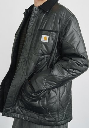 Hombre con chaqueta negra acolchada Carhartt con botones a presión, dos bolsillos delanteros y cuello de pana, manos en los bolsillos inferiores.