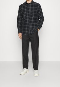Camicia nera con bottoni, decorata con un motivo a griglia bianca, abbinata a pantaloni neri e scarpe bianche. La camicia presenta un colletto e maniche lunghe.