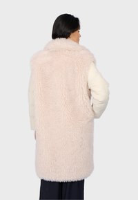 Faux fur gilet in zacht roze, met een fluffy textuur en een brede kraag, gelaagd over een crèmekleurige pufferjack, met een rechte achterkant.