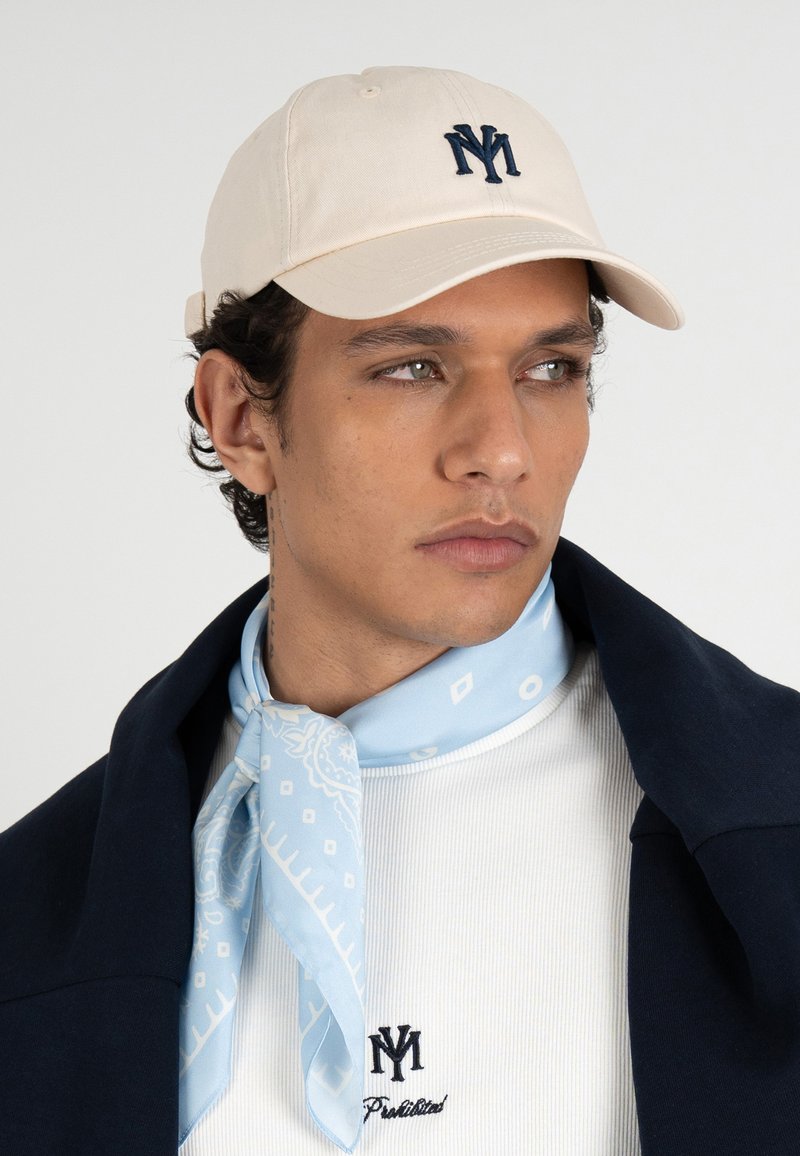 Jeune homme portant une casquette beige avec un logo sombre, un foulard bleu clair à motifs autour du cou, une chemise blanche côtelée avec logo et une veste sombre posée sur les épaules.