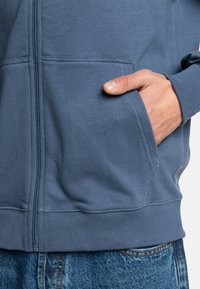 Modrá na zips bunda vyrobená z kombinácie bavlny, so bočnými vreckami, ribbovanými manžetami a hladkou textúrou. Skombinovaná s džínsami.