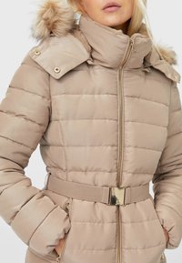 Beige donsjack met capuchon met bontvoering, ritssluiting aan de voorkant, taille met ceintuur, metalen accenten en horizontale doorgestikte structuur. Geschikt voor koud weer.