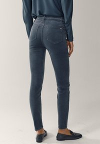 Slim-fit mörkgrå jeans bärs av en stående person, kombinerat med svarta loafers och en långärmad mörk topp, visad från baksidan.