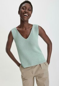 Lichtgroen, gebreid mouwloos topje met een diepe V-hals. Heeft een gestructureerd patroon en een rechte zoom. Gecombineerd met beige broeken.