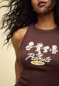 Hnědý žebrovaný top na ramínka s kreslenými postavičkami z Peanuts hrajícími na hudební nástroje. Text zní "Peanuts RECORDS" v bílé a žluté barvě.
