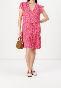 Rosa geblümtes Kleid mit gerafften Kappärmeln, V-Ausschnitt und gestuftem Saum. Kombiniert mit einer braunen Handtasche und hellen flachen Schuhen.