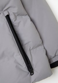 Chaqueta acolchada gris con una textura suave, que presenta un cierre negro visible y un bolsillo. Acabado con gruesos puños negros.