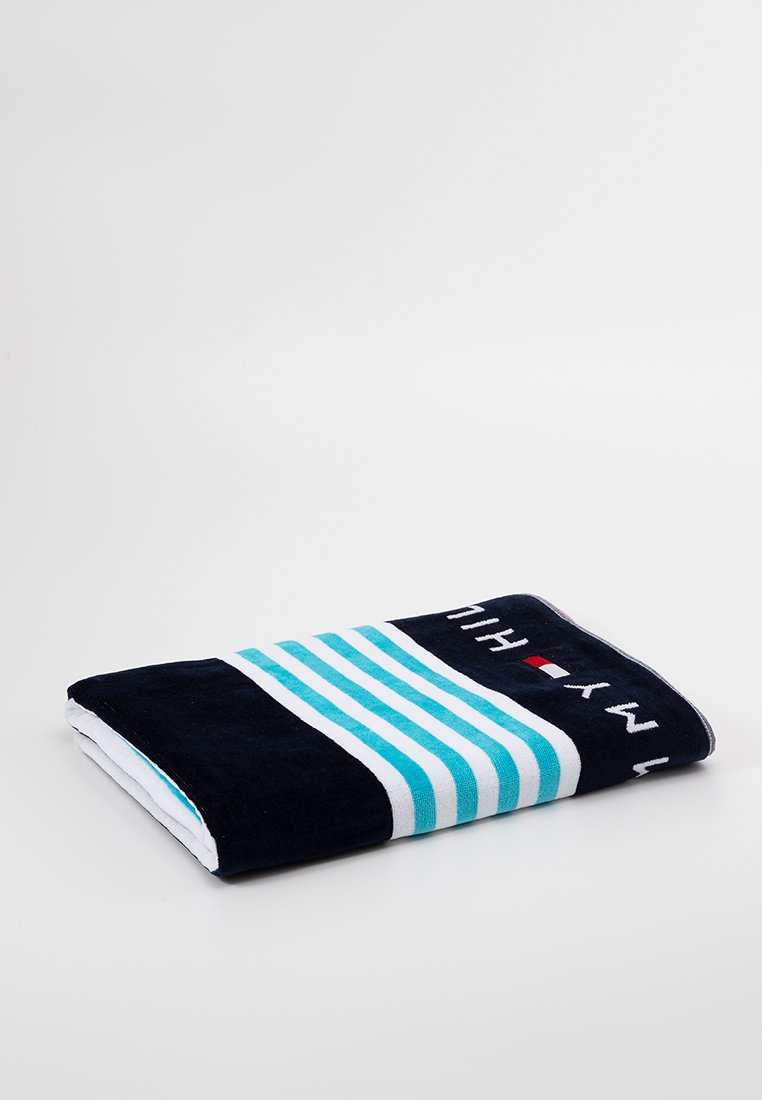 Toalla de color azul marino con rayas turquesa horizontales y detalles en blanco. Presenta texto bordado y un pequeño logo. Tela suave y esponjosa.