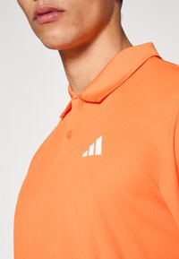 Polo naranja hecho de tela suave, con cuello clásico, tapeta de dos botones y un logo blanco con tres barras en el pecho izquierdo.