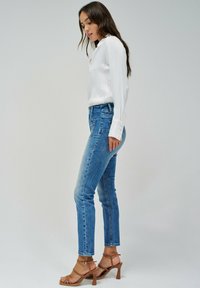 Blusa blanca ligera con cuello, combinada con jeans de mezclilla azul de cintura alta. Los jeans tienen un aspecto desgastado y están estilizados con sandalias de tacón marrón.