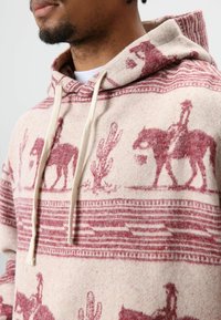 Sweat à capuche en crème avec un design western rouge ; présente des cactus et des cavaliers. Texture douce avec cordons de serrage et accents à motifs.