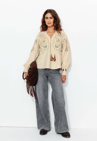 Blouse beige brodée avec des motifs floraux, associée à un jean large gris. Sac en suède marron avec des franges, bottes sombres, design texturé.