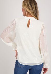 Witte blouse met doorschijnende puffmouwen, bloemenborduurwerk en een keyhole-detail aan de achterkant. Gemaakt van gladde stof met een losse pasvorm, gecombineerd met jeans.