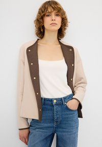 Giacca trapuntata con esterno beige e fodera marrone, dotata di bottoni a pressione dorati. Abbinata a una maglietta bianca e jeans blu a vita alta.