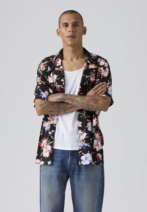Homme aux cheveux courts et tatouages croisant les bras, portant une chemise noire à fleurs sur un débardeur blanc et un jean bleu, sur un fond uni.