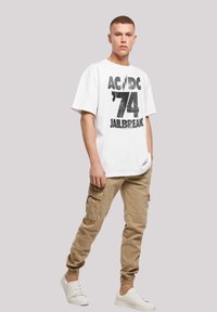 White crew-neck t-shirt with black AC/DC '74 Jailbreak print, paired with beige cargo pants and white sneakers. Loose fit, smooth fabric texture.
