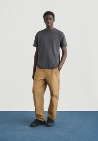 Γκρι βαμβακερό μπλουζάκι με μικρό χρυσό λογότυπο, συνδυασμένο με μπεζ υφασμάτινο παντελόνι και μαύρα αθλητικά παπούτσια. Στέκεται πάνω σε μπλε χαλί μπροστά σε λευκό φόντο.