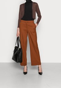 Kobieta w brązowej krótkiej kardiganie, czarnym golfie, rudych spodniach o szerokich nogawkach, czarnych szpilkach, trzymająca czarną skórzaną torbę, pozująca na tle białej ściany.