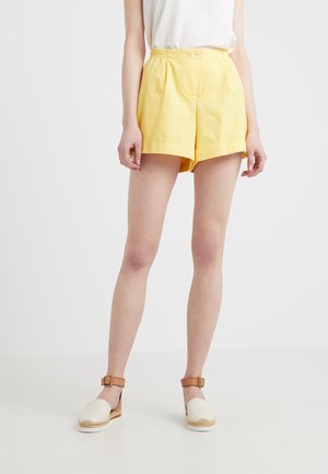 Pantalones cortos de algodón amarillos con un ajuste relajado, detalle de pliegues y doble botón en la parte delantera. Combinados con alpargatas de plataforma blancas que tienen una correa marrón.
