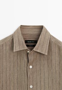 Chemise en lin beige boutonnée avec de fines rayures verticales, col classique et boutons blancs, étiquetée Massimo Dutti.