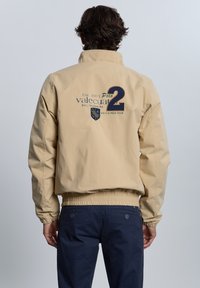 Chaqueta bomber beige con cintura y puños elásticos. Presenta un logo con "Polo 2" y un diseño de blasón en la parte posterior. Combinada con pantalones navy.