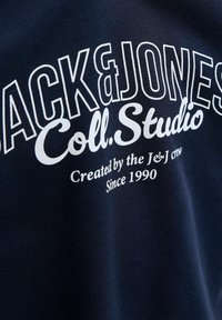 Dunkelblauer Stoff mit weißem Text, der "JACK&JONES Coll. Studio Created by the Je&J crew Since 1990" in fetter und kursiver Schrift liest.