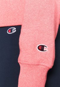 Sudadera con una mezcla de tela rosa suave y azul marino, puños acanalados y logo bordado en rojo y blanco en ambas mangas.