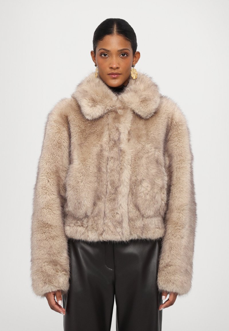 Chaqueta corta de piel sintética en beige claro con un amplio cuello, textura suave y mangas largas. Combinada con pantalones de cuero negros.