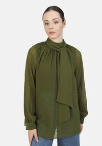 Olive green blouse made of lightweight, textured fabric with a tied neck and puffed sleeves, featuring small raised dots and a relaxed fit.