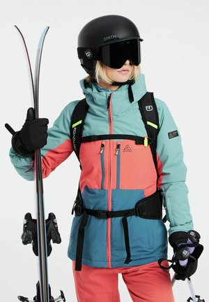 Skibekleidung mit einer türkisfarbenen und korallenfarbenen Jacke im taillierten Design, schwarzen Handschuhen, Helm, Skistöcken und passenden Skihosen.