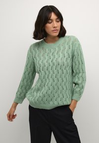 Light green knitted sweater with a loose fit, featuring a textured wave pattern and 3/4 length sleeves. Ribbed neckline and hem details.