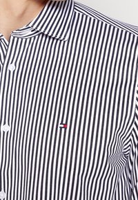 Camicia a righe con bottoni in tessuto blu navy e bianco, con un piccolo logo ricamato in rosso, bianco e blu sul petto.