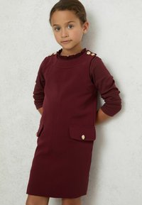 Burgundi színű ruha fodros dekoltárral, arany gombokkal a vállaknál, hosszú ujjakkal és két elülső zsebbel, arany kiemelésekkel.