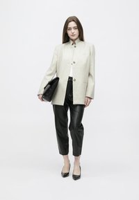 Γυναίκα που φοράει μπεζ παλτό με κουμπιά, μαύρο δερμάτινο παντελόνι cropped, μαύρες μυτερές γόβες, κρατώντας μια μεγάλη μαύρη τσάντα φάκελο.