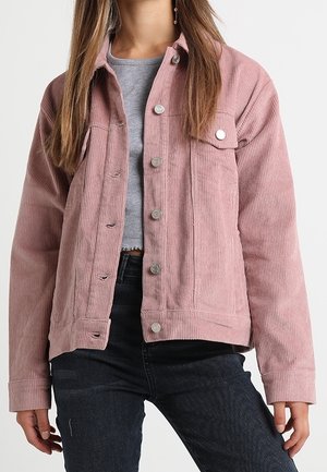 Kvinde iført en støvet rosa fløjlsjakke over en grå kort t-shirt og mørke jeans mod en ensfarvet lys baggrund.