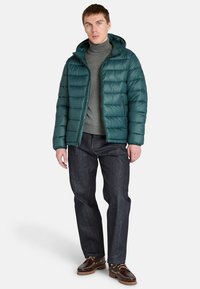 Muž stojí v tyrkysové péřové bundě, šedém roláku a tmavých džínách, má na sobě hnědé boty. Má krátké vlasy a neutrální výraz.