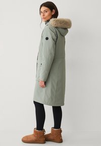 Lichtgroen waterdicht jack met een met bont gevoerde capuchon, zijzakken en zwarte logo-details, gedragen over zwarte leggings en bruine laarzen.