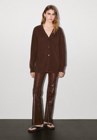 Burgundy stickad cardigan med v-ringning och knappar, kombinerad med åtsittande bruna läderbyxor och svarta flip-flops, modell som står mot en enkel bakgrund.