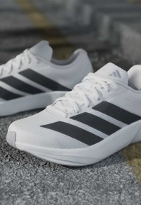 Baskets blanches de sport avec un logo à trois bandes noires, lacées, posées sur une surface pavée extérieure texturée.