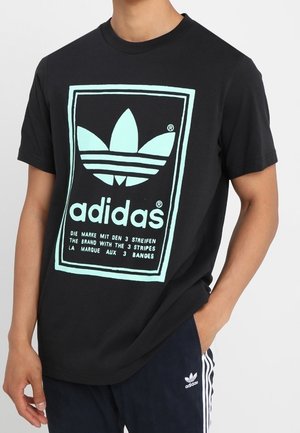 Czarna bawełniana koszulka z dużym miętowozielonym logo Adidas w prostokątnym obramowaniu, z tekstem w kilku językach poniżej.
