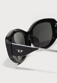 Black oversized sunglasses with a glossy finish, angular frames, and dark lenses. Features metallic accent on the temple.