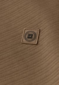 Tessuto marrone dalla texture con un motivo lineare in rilievo, dotato di un'etichetta rettangolare beige embossata con un logo nero al centro.