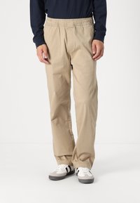 Beige, elastic-waist trousers with a straight-leg design. Made from a soft, lightweight fabric. Worn with white and grey sneakers.
